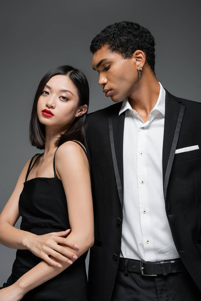 asian woman in black strap dress posing near young african american man in elegant blazer isolated on grey - 写真・画像