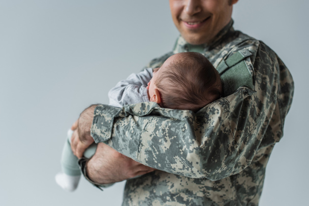 cheerful serviceman in camouflage uniform holding newborn son isolated on grey  - Фото, зображення