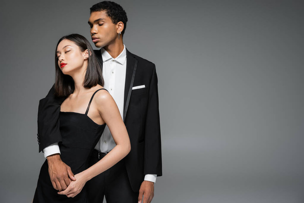 stylish african american man in elegant suit embracing asian woman posing with closed eyes isolated on grey - Фото, изображение