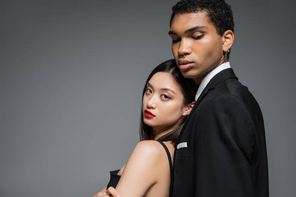 charming asian woman with red lips leaning on african american man in black blazer isolated on grey - Zdjęcie, obraz