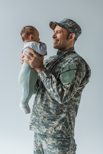 positive army soldier in uniform holding newborn son in arms isolated on grey  - Foto, Imagem