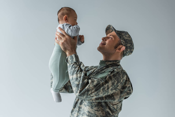 Ευτυχισμένος στρατιώτης με στολή που κρατάει το αγοράκι στην αγκαλιά του απομονωμένο στο γκρι.  - Φωτογραφία, εικόνα