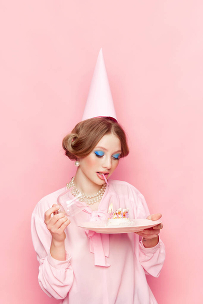 Stylish, beautiful, young girl with makeup opening her birthday cake against pink studio background. Make your wish. Food pop art photography. Concept of retro style, creative vision, imagination. - Foto, Imagen