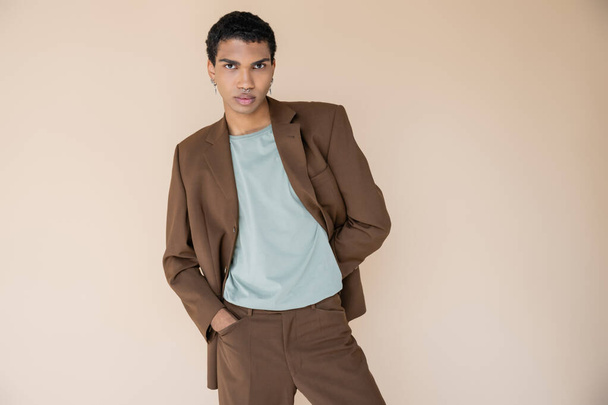 young african american man in brown suit standing with hand in pocket and looking at camera isolated on beige - Photo, Image
