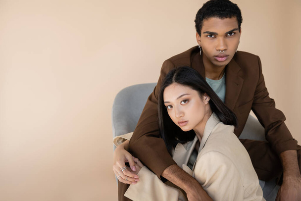 african american man in brown blazer embracing brunette asian woman and looking at camera isolated on beige - Photo, Image