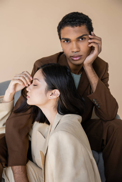 stylish african american man in brown suit looking at camera near sensual asian woman with closed eyes isolated on beige - 写真・画像