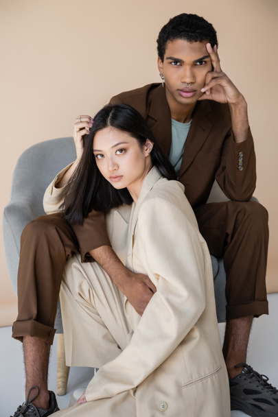 fashionable interracial models in trendy pantsuits looking at camera near armchair on beige background - Фото, изображение