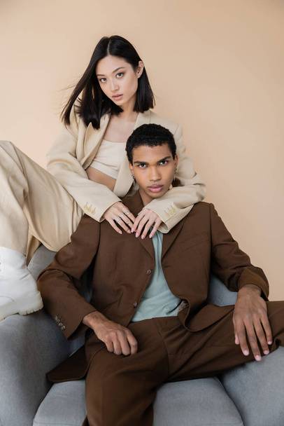 sensual asian woman embracing african american man in trendy suit sitting in armchair isolated on beige - 写真・画像