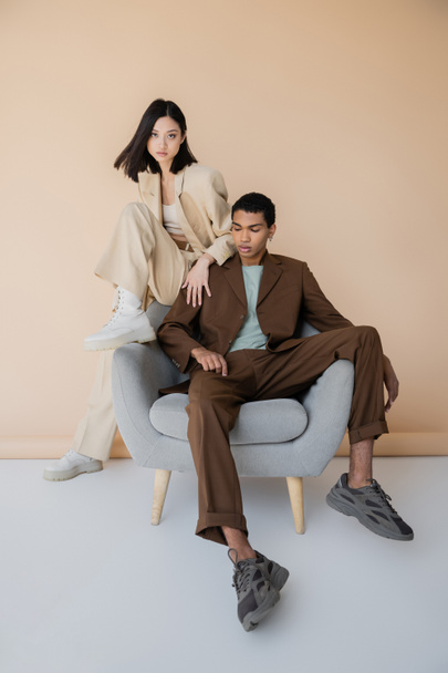 full length of young interracial couple in fashionable pantsuits posing on armchair on beige background - Photo, Image
