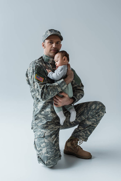 Soldado estadounidense en uniforme sosteniendo en brazos al recién nacido y mirando a la cámara en gris - Foto, imagen