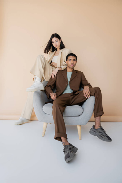 longitud completa de pareja interracial en elegantes trajes de pantalón posando cerca de sillón sobre fondo beige - Foto, imagen