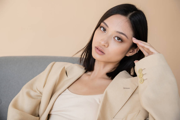 young asian woman in blazer holding hand near face and looking at camera on beige background - Фото, зображення