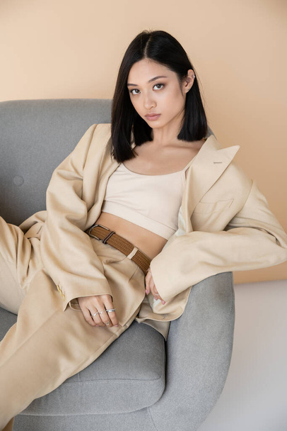 sensual asian woman in stylish pantsuit sitting in armchair and looking at camera on beige background - Photo, Image