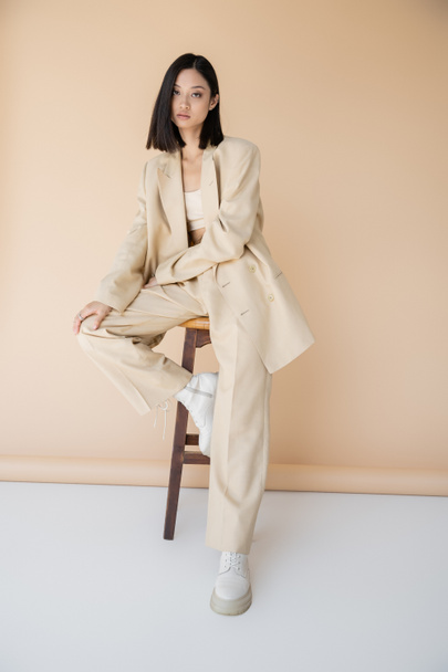 brunette aasialainen nainen tyylikäs housupuku ja saappaat istuu jakkaralla beige tausta - Valokuva, kuva