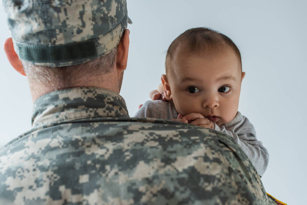 father in military uniform and cap hugging newborn son isolated on grey  - Photo, Image