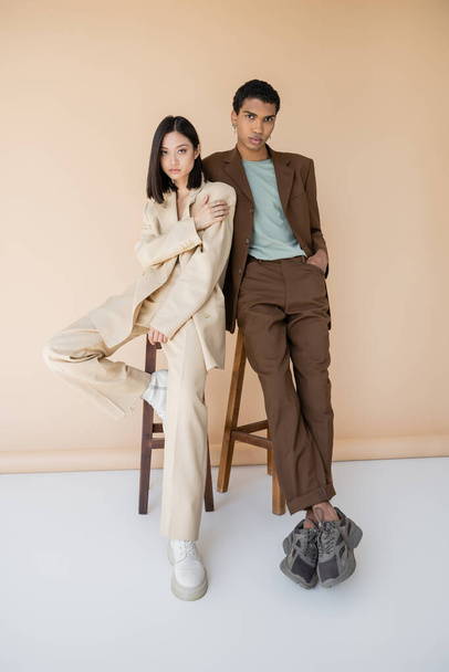 piena lunghezza di giovane coppia multietnica in abiti eleganti posa vicino sgabelli su sfondo beige - Foto, immagini