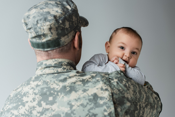 Soldat in Militäruniform und Mütze umarmt neugeborenen Sohn isoliert auf grau  - Foto, Bild