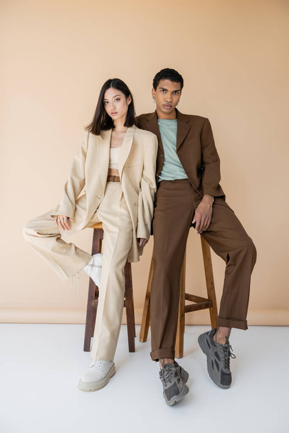 full length of multiethnic couple in stylish pantsuits posing near stools on beige background - 写真・画像