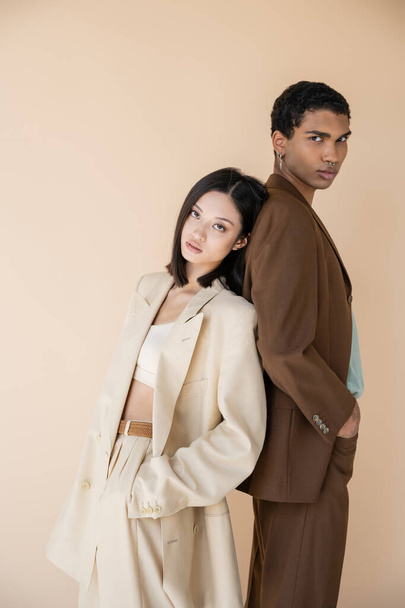trendiges gemischtrassiges Paar in Anzügen, Rücken an Rücken stehend, die Hände in Taschen isoliert auf beige - Foto, Bild
