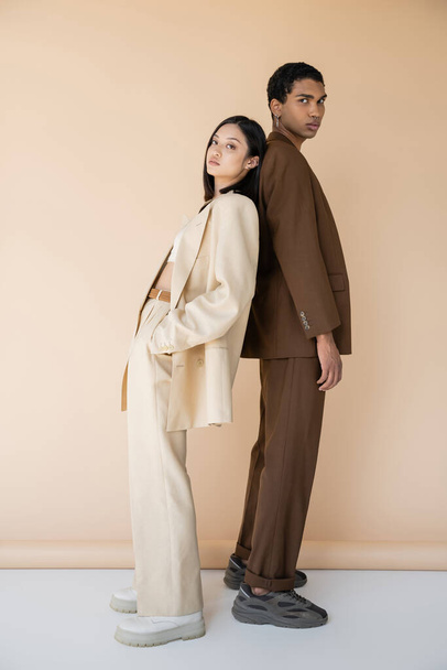 full length of asian woman and african american man in trendy pantsuits standing back to back on beige background - Fotografie, Obrázek