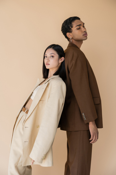 young asian woman in pantsuit standing back to back with trendy african american man and looking at camera isolated on beige - Foto, imagen