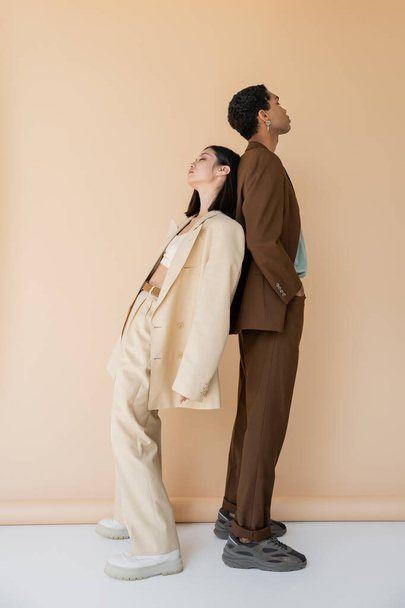 full length of interracial couple in stylish pantsuits posing back to back on beige background - Foto, Imagen
