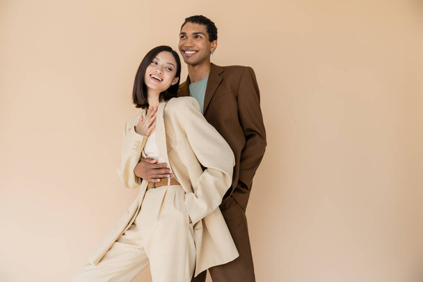 positive african american man embracing excited asian woman in ivory suit isolated on beige - Fotó, kép
