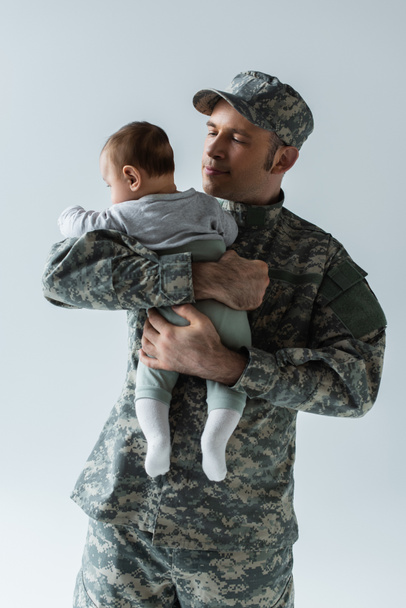 proud serviceman in military uniform and cap hugging infant son isolated on grey  - Valokuva, kuva