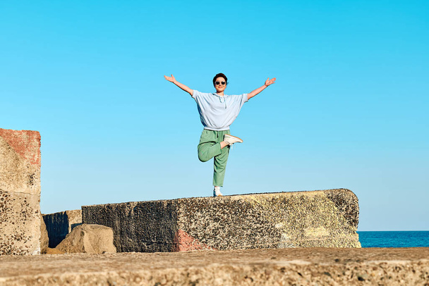 Szczęśliwa turystka ciesząca się wolnością podczas jogi na wielkim kamieniu na molo z błękitnym niebem w tle. - Zdjęcie, obraz