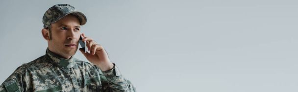 Soldat in Mütze und Armeeuniform unterhält sich auf Smartphone isoliert auf Grau, Banner  - Foto, Bild