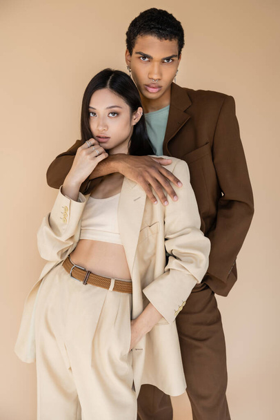 trendy african american man embracing asian woman in pantsuit and looking at camera isolated on beige - Foto, afbeelding