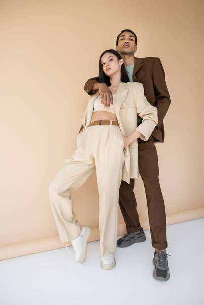 full length of african american man hugging trendy asian model in ivory pantsuit on beige background - Photo, Image