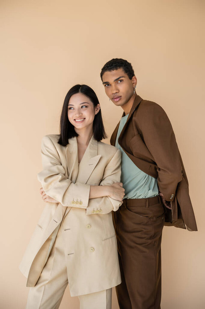 cheerful asian woman posing with folded arms near african american man in brown suit isolated on beige - Foto, afbeelding