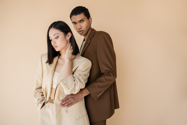 young african american man in brown blazer embracing brunette asian woman and looking at camera isolated on beige - Foto, Bild