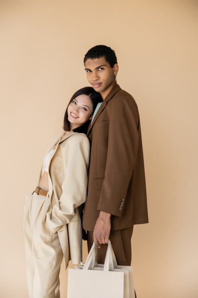 elegante donna asiatica sorridente vicino afro americano uomo con shopping bags isolato su beige - Foto, immagini