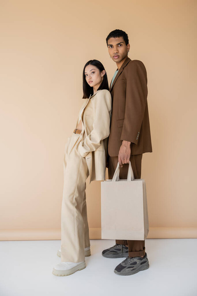 full length of asian woman in pantsuit posing with hand in pocket near african american guy with shopping bags on beige background - Фото, изображение
