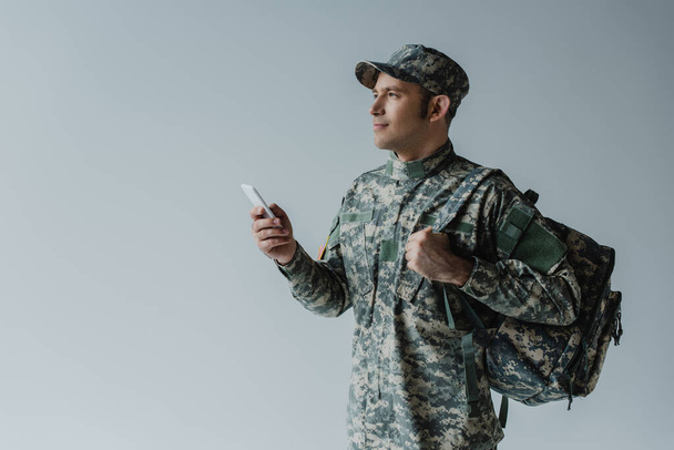 serviceman in cap and army uniform standing with backpack and holding smartphone isolated on grey  - Foto, Imagem