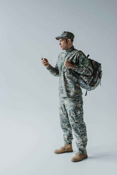 militaire en casquette et uniforme de l'armée debout avec sac à dos et smartphone sur gris  - Photo, image