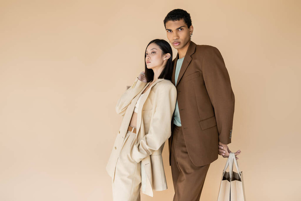 african american man in brown suit posing with shopping bags near trendy asian model isolated on beige - Foto, immagini