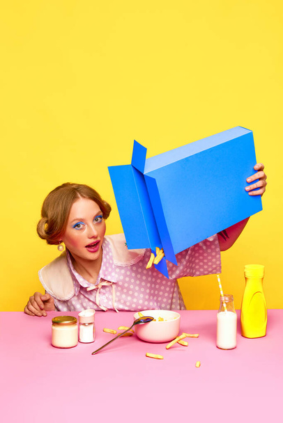 Fast food lover. Pretty young girl with bright makeup pouring fries from box against yellow studio background. Food pop art photography. Concept of retro style, creative vision. Complementary colors - Photo, image