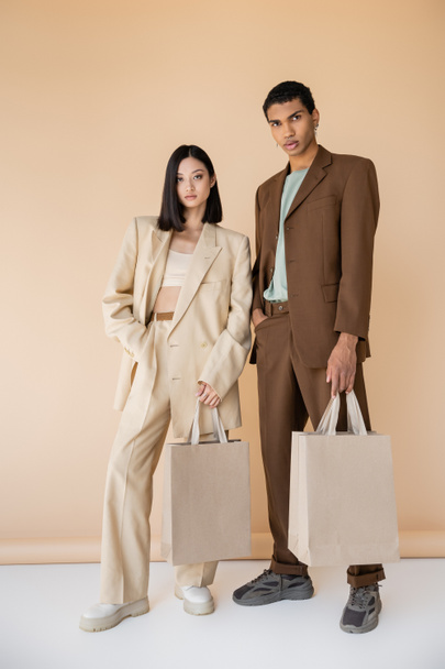 longitud completa de pareja interracial en trajes de moda posando con bolsas de compras sobre fondo beige - Foto, Imagen