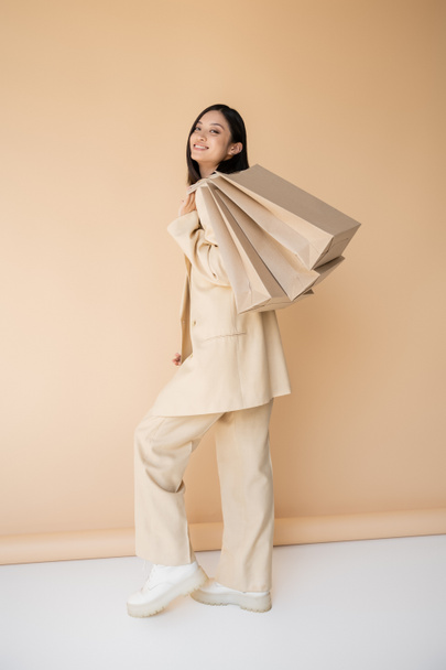 volledige lengte van vrolijke aziatische vrouw in stijlvolle broekpak poseren met boodschappentassen op beige achtergrond - Foto, afbeelding