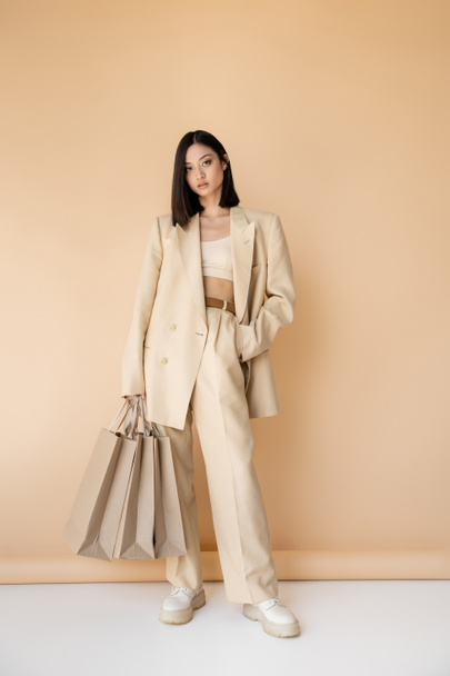 Tam boy esmer Asyalı şık takım elbiseli kadın bej arka planda alışveriş torbalarıyla ayakta duruyor. - Fotoğraf, Görsel