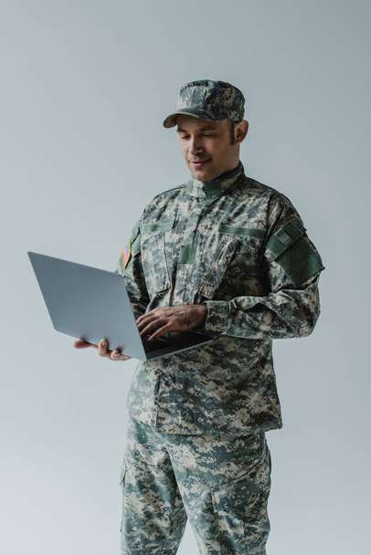 good looking soldier in army uniform and cap using laptop isolated on grey  - 写真・画像