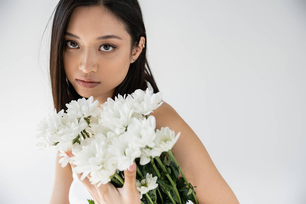 portrait of sensual asian woman with natural makeup looking at camera near bouquet of white flowers isolated on grey - Фото, изображение