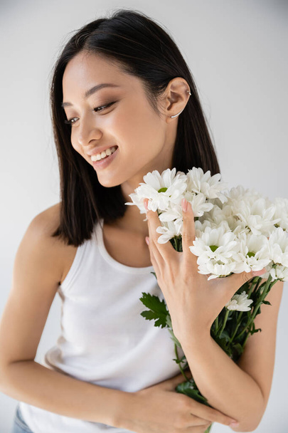 morena mujer asiática con pendiente de puño sonriendo cerca de flores blancas aisladas en gris - Foto, imagen
