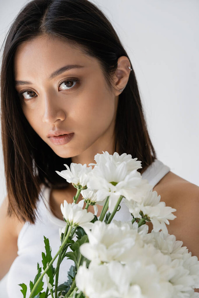 portrait of sensual asian woman with brunette hair and natural makeup near white flowers isolated on grey - Zdjęcie, obraz