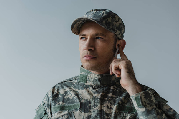 soldier in uniform and cap adjusting wireless earphone isolated on grey  - Фото, изображение