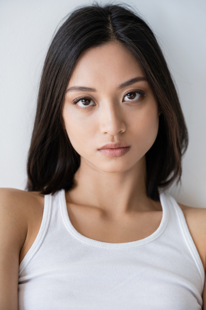 portret van brunette aziatische vrouw in wit tank top kijken naar camera geïsoleerd op grijs - Foto, afbeelding
