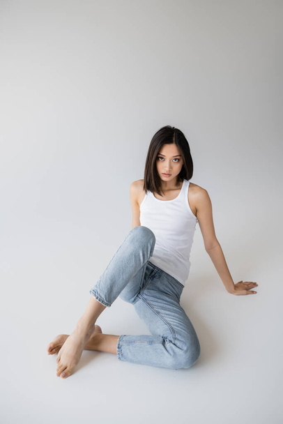full length of barefoot asian woman posing in white tank top and blue jeans on grey background - Foto, Imagem